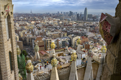 Spain, Catalonia, Barcelona, Eixample district, Sagrada Familia basilica by Catalan modernist architect Antoni Gaudi, listed as a UNESCO World Heritage Site, peaks topped with mosaics in the shape of fruits surrounding the construction site on the roof of the nave at the rear of the future facade of the Glory, the Torre Agbar by architect Jean Nouvel in the background